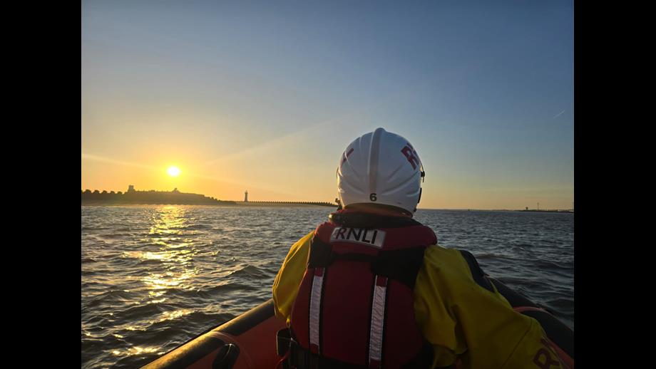 RNLI volunteer crew can be seen centre of the photo, helming the lifeboat. Beyond, Fort Perch Rock is visible with a stunning sunset sky behind.
