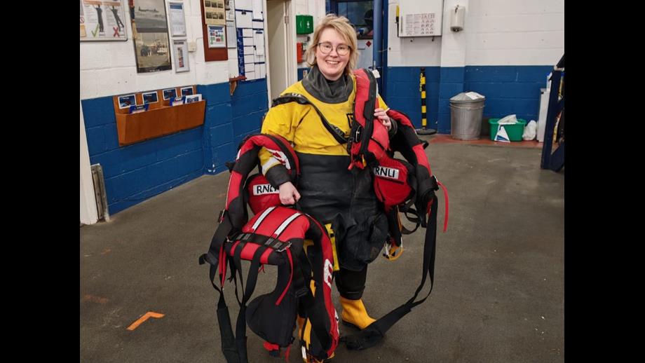A volunteer crew member stands holding multiple life jackets, inside New Brighton boathouse, post-exercise.
