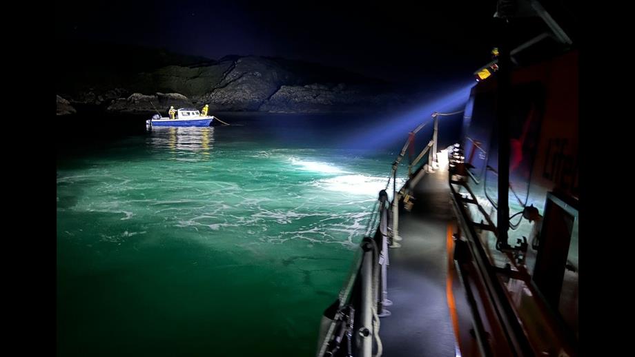 A searchlight from the Oban Lifeboat illuminates a small blue-hulled motorboat in the darkness with two lifeboat crew members visible onboard