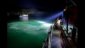 A searchlight from the Oban Lifeboat illuminates a small blue-hulled motorboat in the darkness with two lifeboat crew members visible onboard