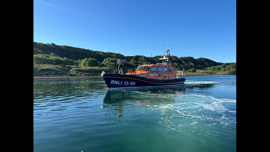 The Oban lifeboat in the morning sunshine, the calm waters frothed up as the lifeboat moves astern from the yacht.
