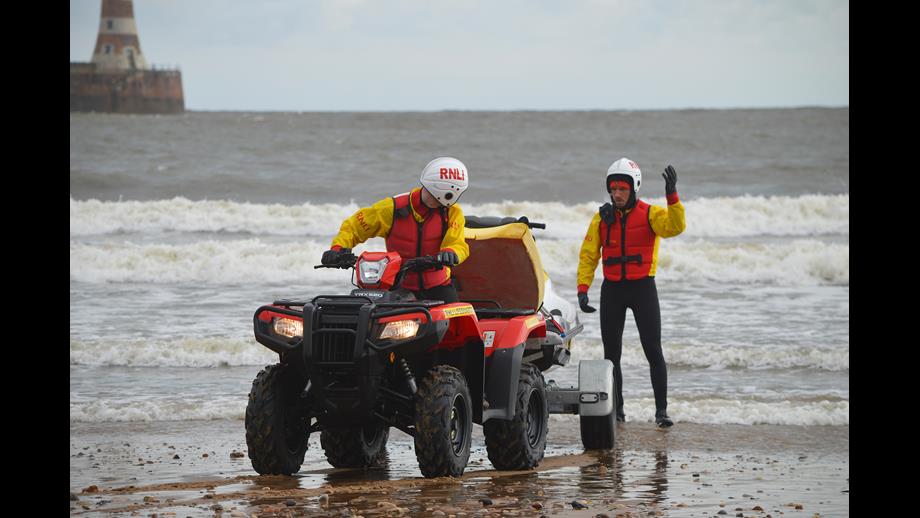 Lifeguard Supervisor Training at Roker Sunderland two Lifeguards in full RNLI lifeguard kit with RNLI quad bike on shore line. Lighthouse and sea in background
