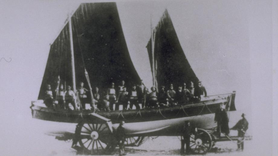 Cromer lifeboat Louisa Heartwell on her carriage. The crew are wearing lifejackets and holding oars aloft. 