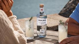 Two people sit at a wooden table, each with a drink and a bottle of Four Seas Salcombe Gin on the table 