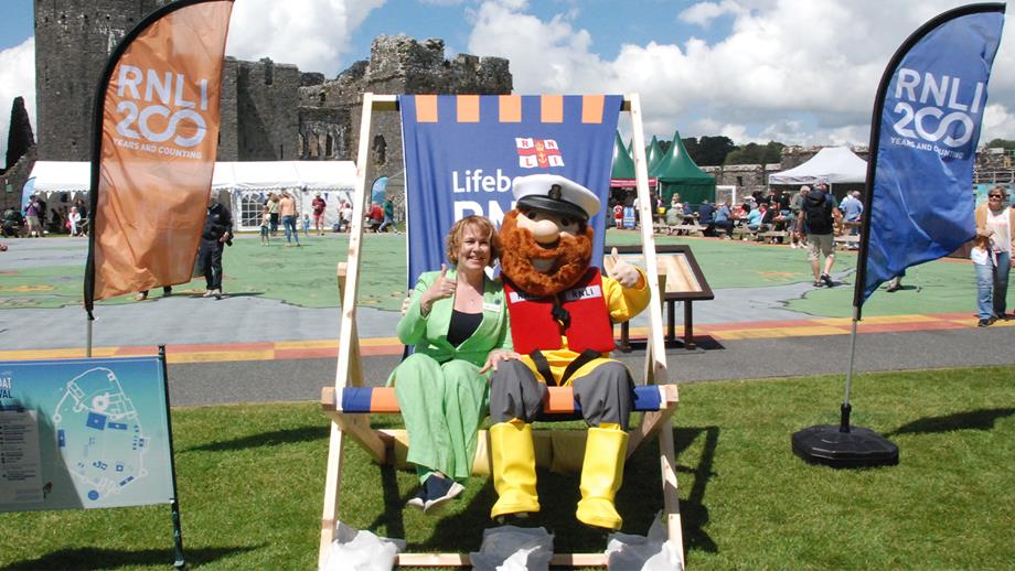 Two people in a giant RNLI deckchair at an RNLI lifeboat festival in 2024 with Pembroke Castle in the background. One is dressed in an RNLI Stormy Stan costume. 