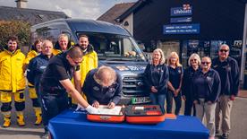 An RNLI volunteer signing the RNLI 200th anniversary scroll on a table in front of RNLI Moelfre Seawatch Centre with other volunteers behind and an RNLI Ford E-Transit van 