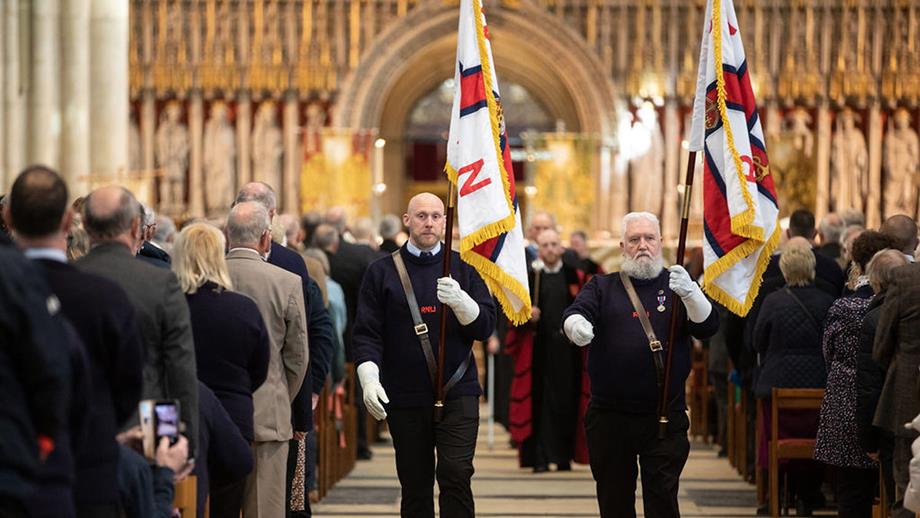 Inside York Minster Cathedral during the RNLI’s Thanksgiving Service on 20 April 2024.  Two RNLI representatives are walking down the aisle, towards the camera, holding RNLI flags.