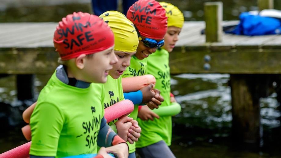 Four children of different ethnicities wade into the water together beside a jetty at a lake, linking arms and holding tube floats under their armpits. They wear red or yellow swim hats, neon green Swim Safe T-shirts and happy smiles on their faces!