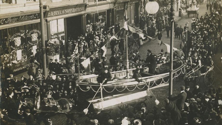 Black and white image of lifeboat being pulled through crowded street, crew collecting donations from crowd