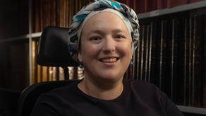 A female heritage volunteer wearing a headscarf and using wheelchair is pictured smiling and facing the camera. She is in a library with books in the background. 