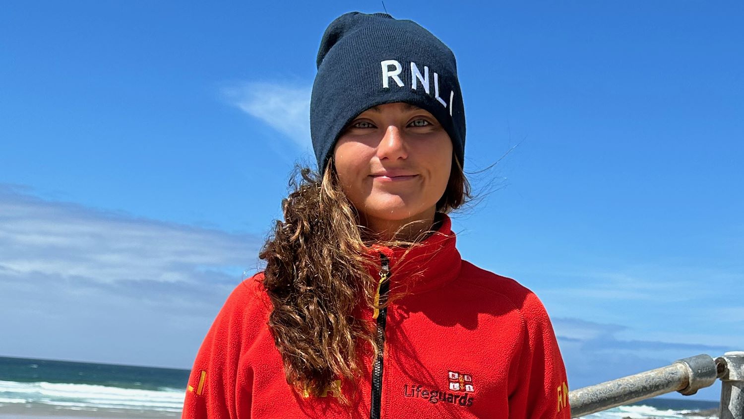 RNLI Lifeguard Elise Irish smiling, with blue skies and the sea in the background.