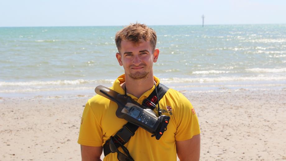 RNLI Senior Lifeguard Zeph Palmer in a lifeguard tshirt smiling at a sandy beach