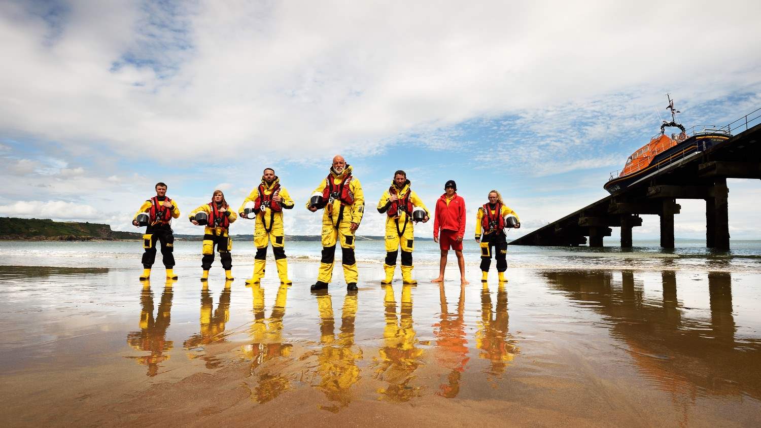 RNLI lifeboat volunteers and an RNLI lifeguard standing on the beach at Tenby with Tenby’s Tamar class all-weather lifeboat on the slipway in the background
