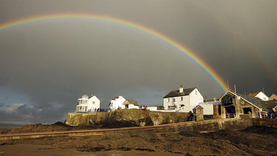 A view from the beach of Appledore Lifeboat Station in north Devon. The tide is out, showing the slipway running down the beach. The sky is grey and cloudy and there is a double rainbow in the sky