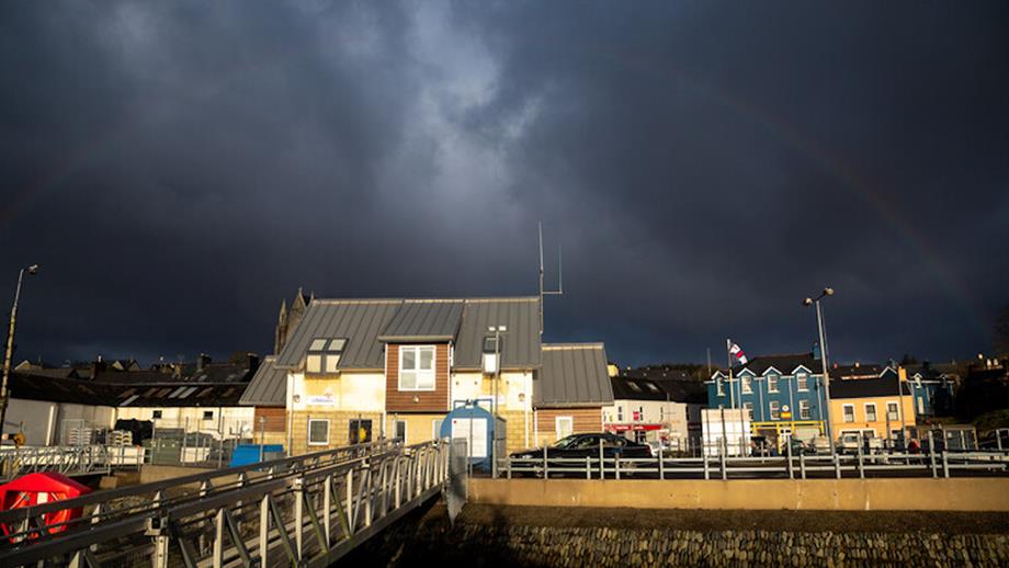 A view of Castletownbere Lifeboat Station in dark overcast weather. The view is from the pontoon, looking up the gangway towards the station. A rainbow can be seen overhead and the sky is moody, cloudy and dark.