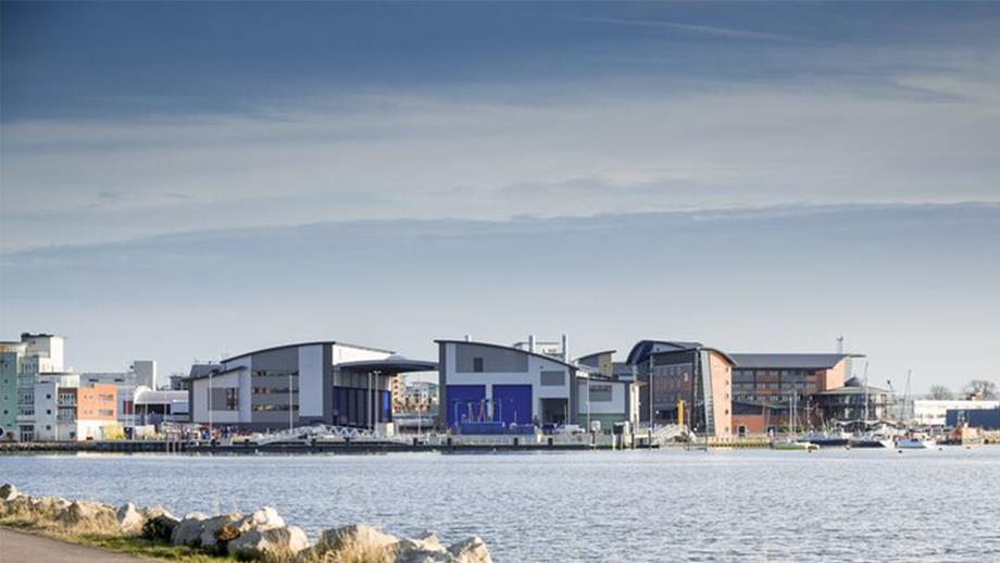 A view across Holes Bay towards the All-weather Lifeboat Centre (ALC) in Poole, Dorset. The photograph is taken from a tarmac path lined by rocks, with the ALC in the background.