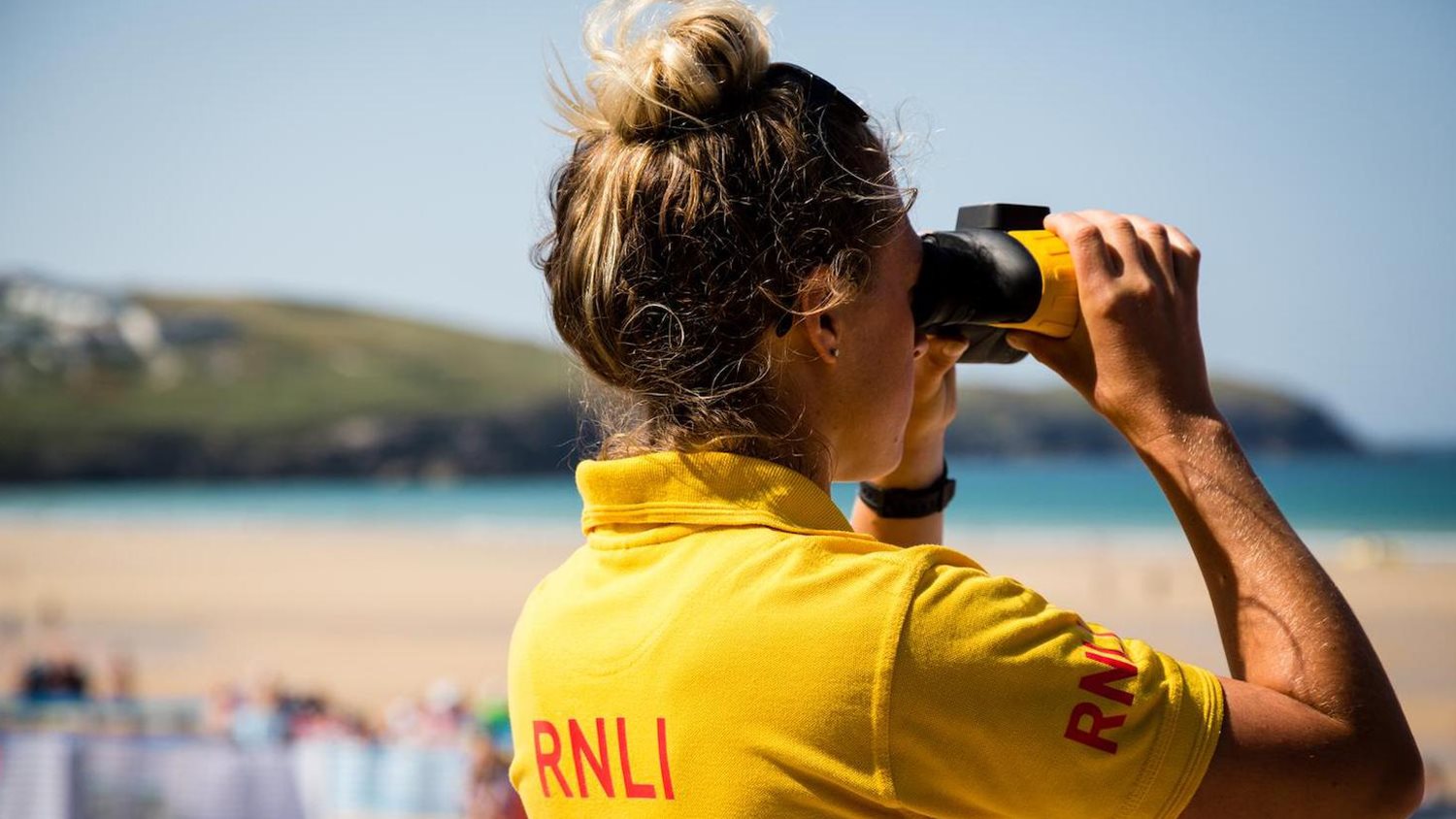 A white lifeguard with highlighted light brown hair in a top bun looks out the the frame through binoculars. They are wearing a yellow RNLI lifeguard polo shirt and the setting is a sandy beach with turquoise water and grass-covered cliffs in the background