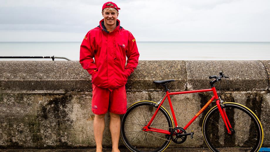 A landscape portrait of a white lifeguard with short blonde hair standing next to a red lifeguard bicycle on a promenade. The lifeguard is wearing lifeguard kit of red shorts, a red waterproof jacket and red baseball cap. The bike is beside them, leaning against a concrete wall with a calm sea in the background and cloudy white sky.