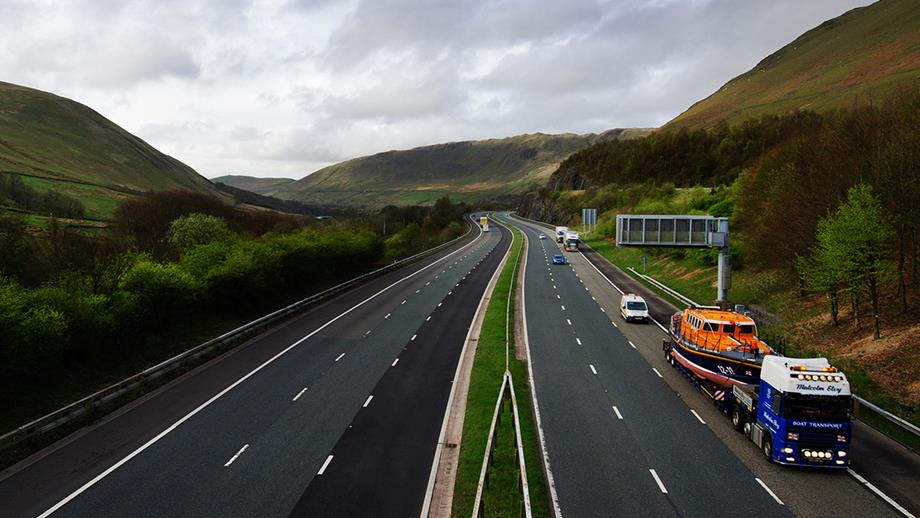 A view from a bridge of a Mersey class lifeboat on a blue and white HGV, in transit along a motorway running through a lush green valley.