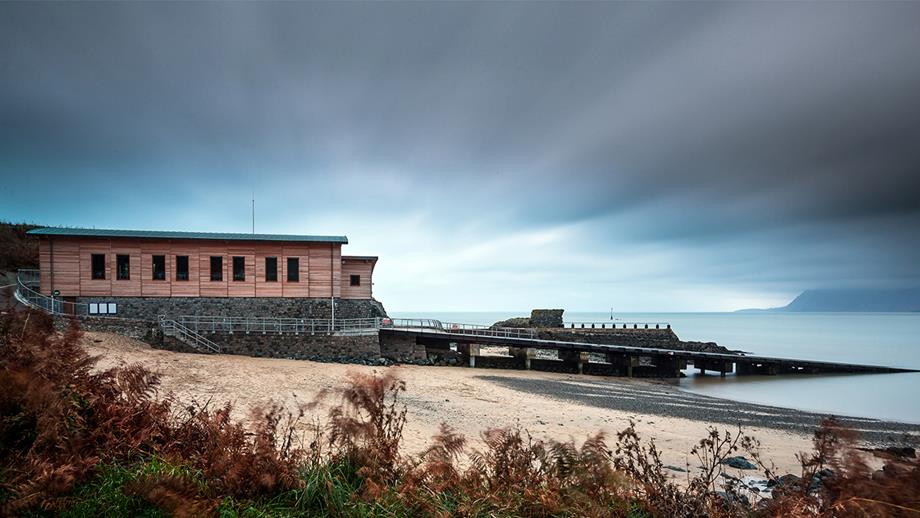 An autumnal view along the coast towards Porthdinllaen Lifeboat Station on the Llyn Peninsula. The lifeboat station is clad in wood and designed for reduced environmental impact. In the foreground is a coastal path with grasses and other wild plants, the station is situated on a yellow sandy beach, and the sea is calm, with mountains visible in the background, beyond the water.