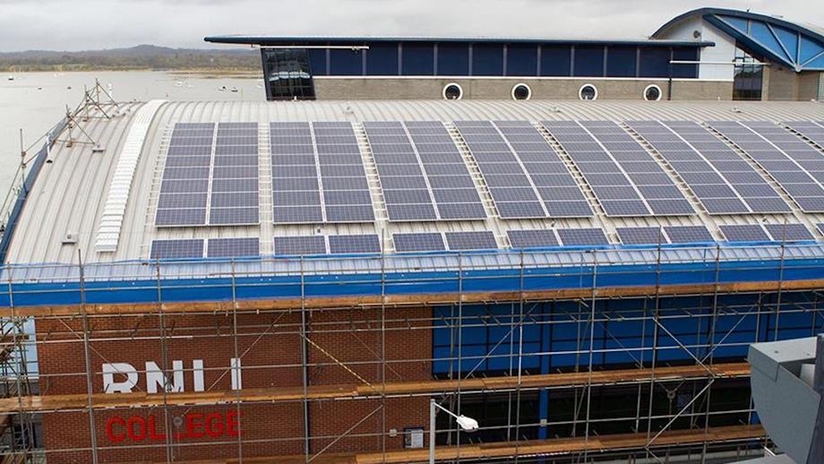 A view of the RNLI’s Sea Survival Centre main building from an elevated aspect, showing the curved roof covered in multiple solar photovoltaic (PV) panels. The building is surrounded by scaffolding and the RNLI Support Centre office building can be seen in the background.