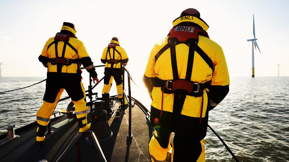 Three crew stand on the bow of the Skegness Shannon class lifeboat, during a search and rescue exercise. A wind farm is in the background.
