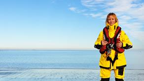 A white lifeboat crew member with brown hair pulled back off their face. They’re looking straight to camera and wearing full lifeboat crew kit – including the new Helly Hansen crew kit, and red lifejacket. Under their arm is a white crew helmet.