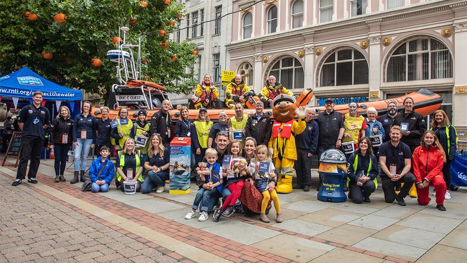 A reconstruction of the world’s first recorded charity street collection, Lifeboat Saturday, in Manchester in October 2021. Crew are standing aboard a lifeboat in the city centre surrounded by RNLI fundraisers of all ages.