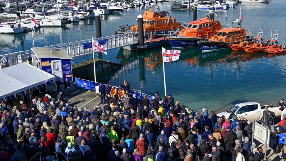 A crowd of people listening to a speech by volunteers, overlooking the harbour, with lifeboats in background.