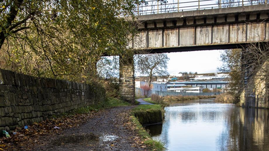 A muddy towpath with the canal to the right, a bridge up ahead and industrial buildings in the distance