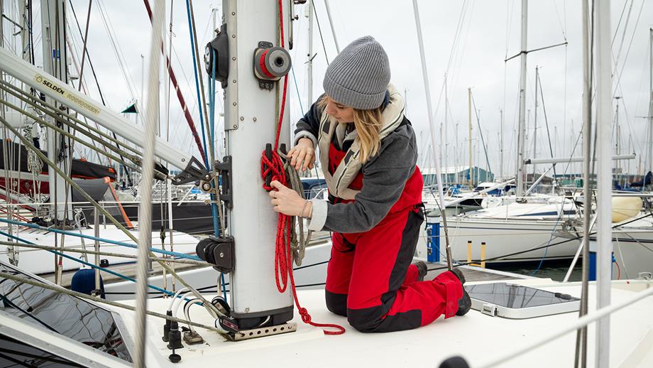 A young woman in a beanie ties a knot on the deck of her boat