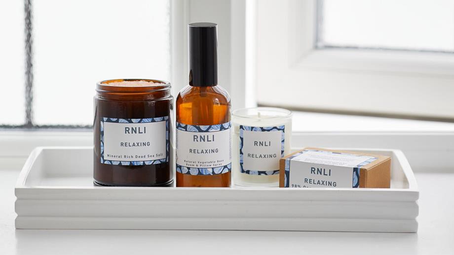 A tray of RNLI-branded beauty products, including bath salts, soap and a candle, sit on a white tray on the windowsill of a bright bathroom