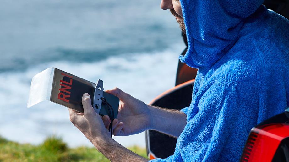 A man in a towelling hooded robe beside the sea is holding their mobile phone in an RNLI-branded waterproof pouch 