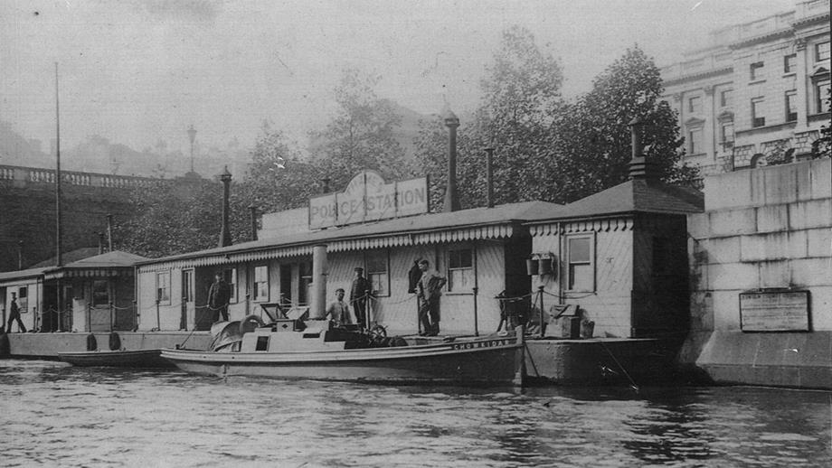 A black and white photograph showing the floating Thames Police Station, with a boat moored in front of it