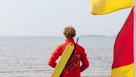 The back of an RNLI lifeguard, looking out to sea. He is wearing a red fleece with a yellow rescue tube over his shoulder and is standing next to a red and yellow flag.