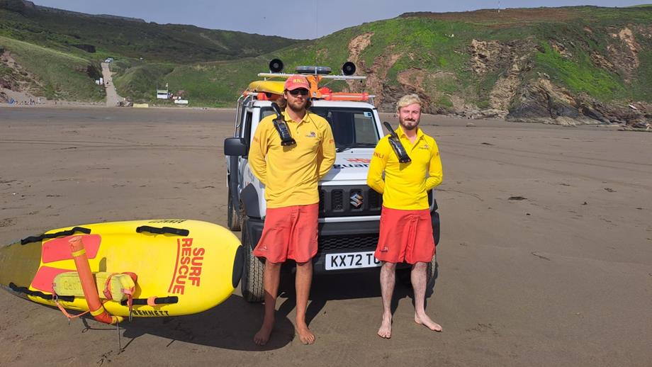 Senior RNLI Lifeguards Rupert Callard (left) and Harry Moir (right) standing on Tregantle Beach in front of the beach patrol vehicle. A rescue board lies on the wet sand next to Rupert.