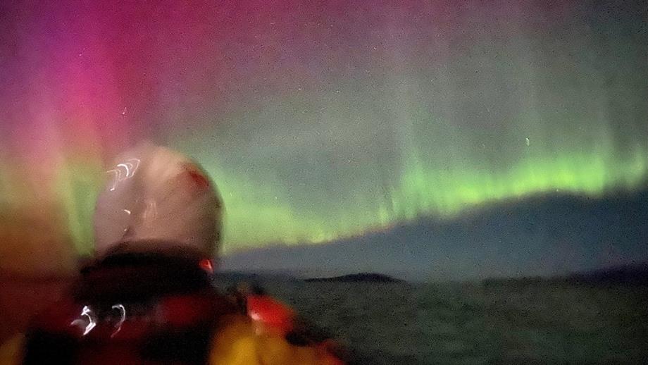 Two crew members in an inshore lifeboat look up at the northern lights, which is making the sky glow pink and green 
