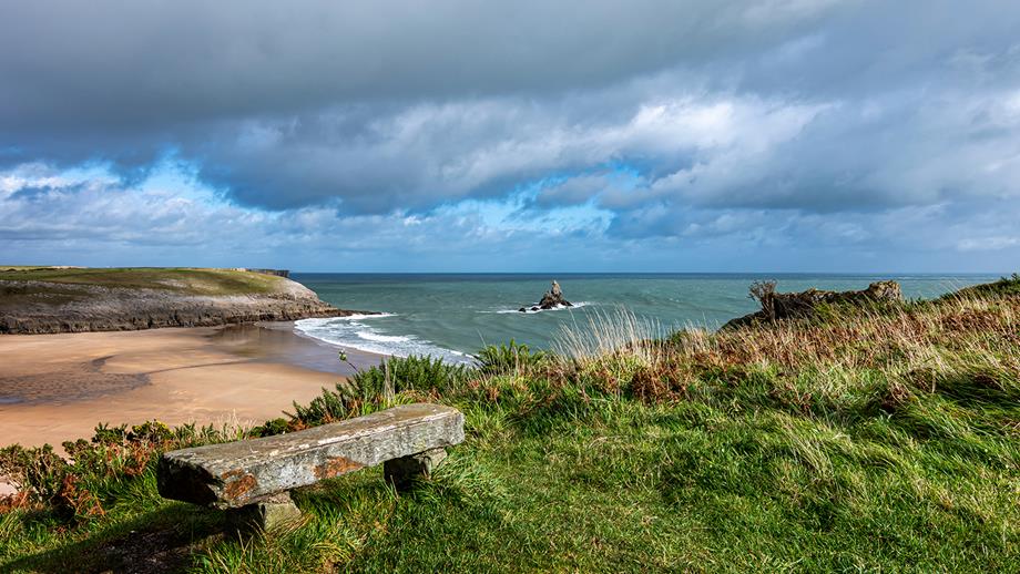 A coastal view with a sandy beach and cliffs and a wooden bench in the foreground