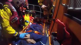 A casualty lies on a stretcher, in an all-weather lifeboat, holding hands with an RNLI crew member 