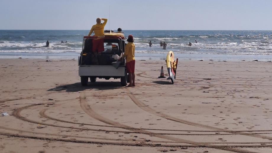Senior RNLI Lifeguards Harry Moir (left) and Rupert Callard (right) keeping watch over swimmers at Tregantle Beach from the beach patrol vehicle. A rescue board lies on the wet sand next to Rupert. The view is of the back of the lifeguards as they look out to sea.