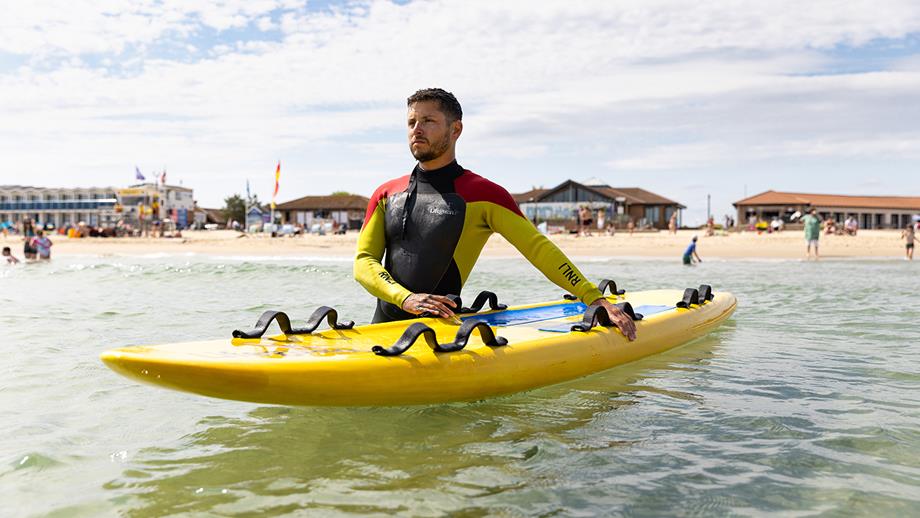 A lifeguard stands waist-deep in water at Sandbanks Beach, Poole, holding a yellow rescue board. 