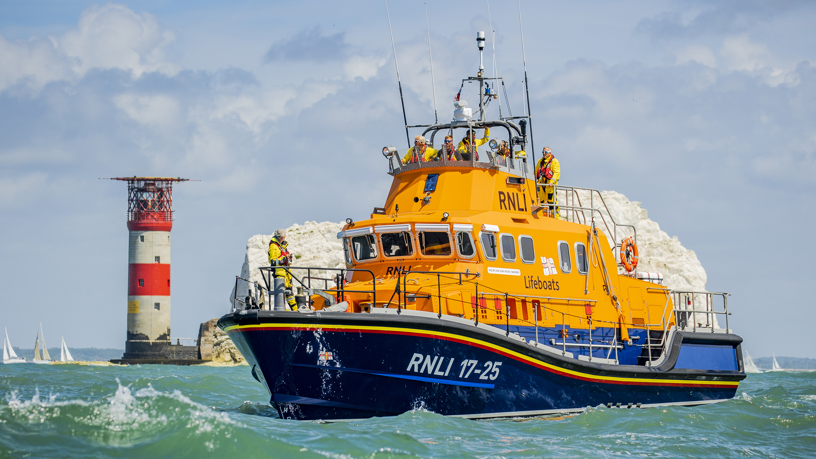 Yarmouth’s Severn class lifeboat passes by The Needles. There is one crew member at the front of the boat on the lower deck, and five crew looking out from the upper deck.