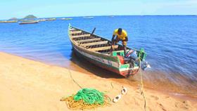 A man stands up in a moored wooden boat on Lake Victoria in Tanzania. There are lots of fishing boats in the background, and yellow and green rope piled on the golden sand in the foreground.