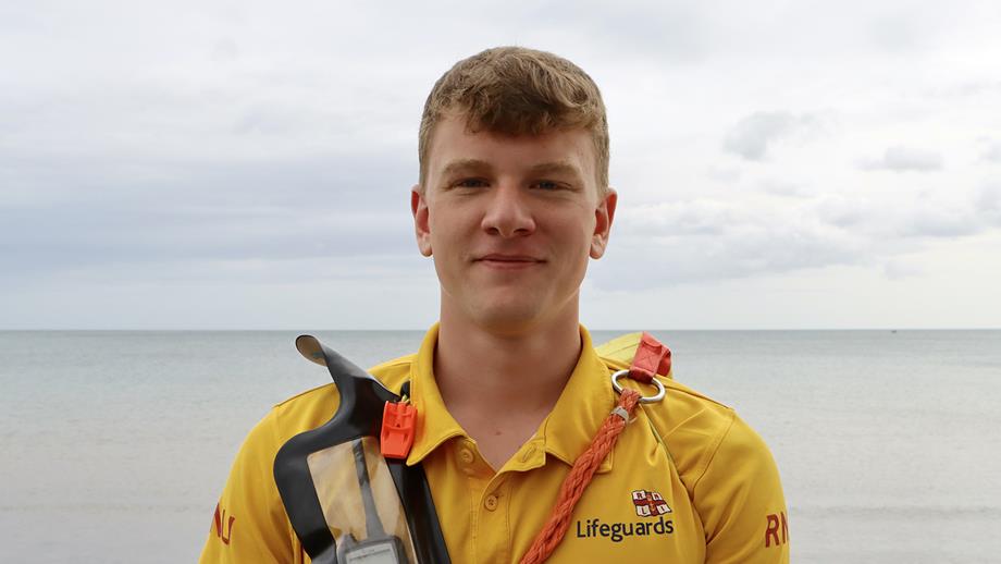 A head and shoulders photo of RNLI Lifeguard Callum Mcdonald, a white man with light brown hair, looking straight to camera with the sea in the distance. He is wearing a yellow lifeguard polo shirt and carries a yellow rescue float over his shoulder and a VHF radio and whistle strapped to his chest.