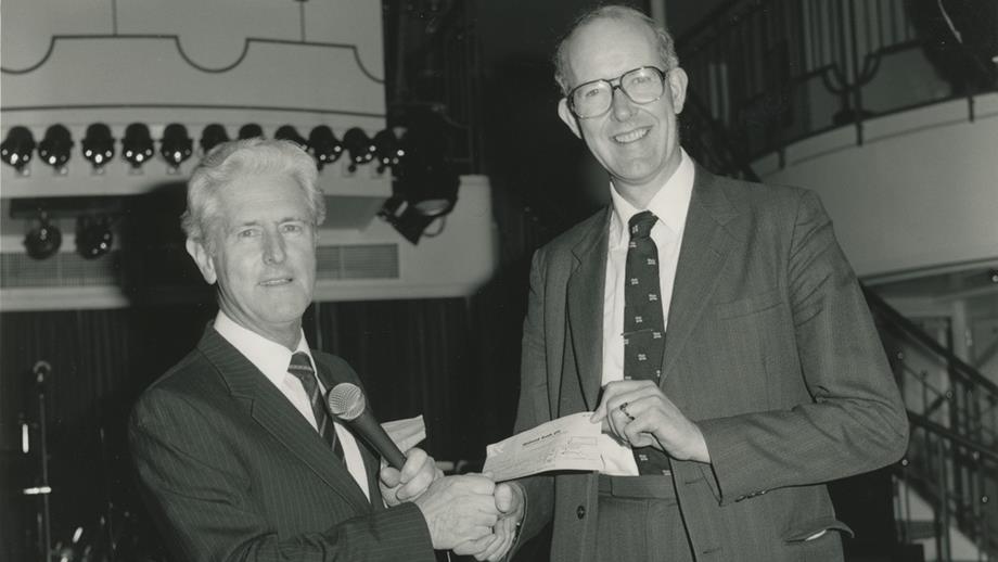 A black and white photograph of two men wearing suits and shaking hands. One of the men is handing a cheque to the other. 