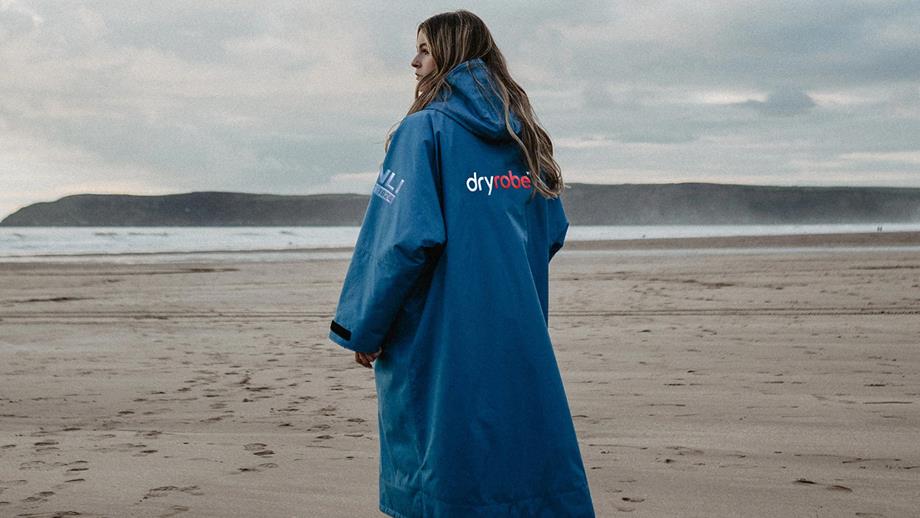 A woman stands on the beach wearing a cosy-looking blue, long-sleeved, hooded changing robe