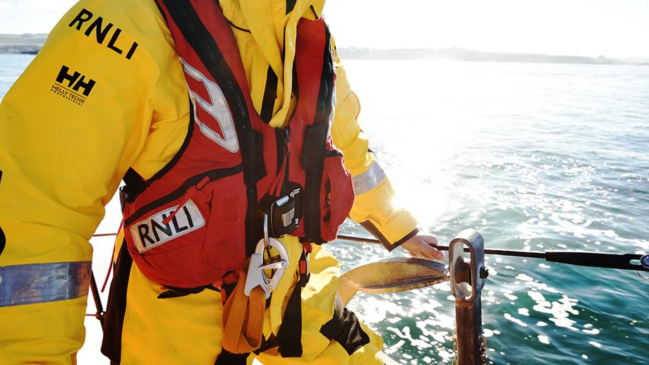 A close-up shot of a crew member wearing Helly Hansen kit onboard an all-weather lifeboat. The crew member’s head is out of shot. The crew member is holding onto the side of the lifeboat. The sun is shining on the water.
