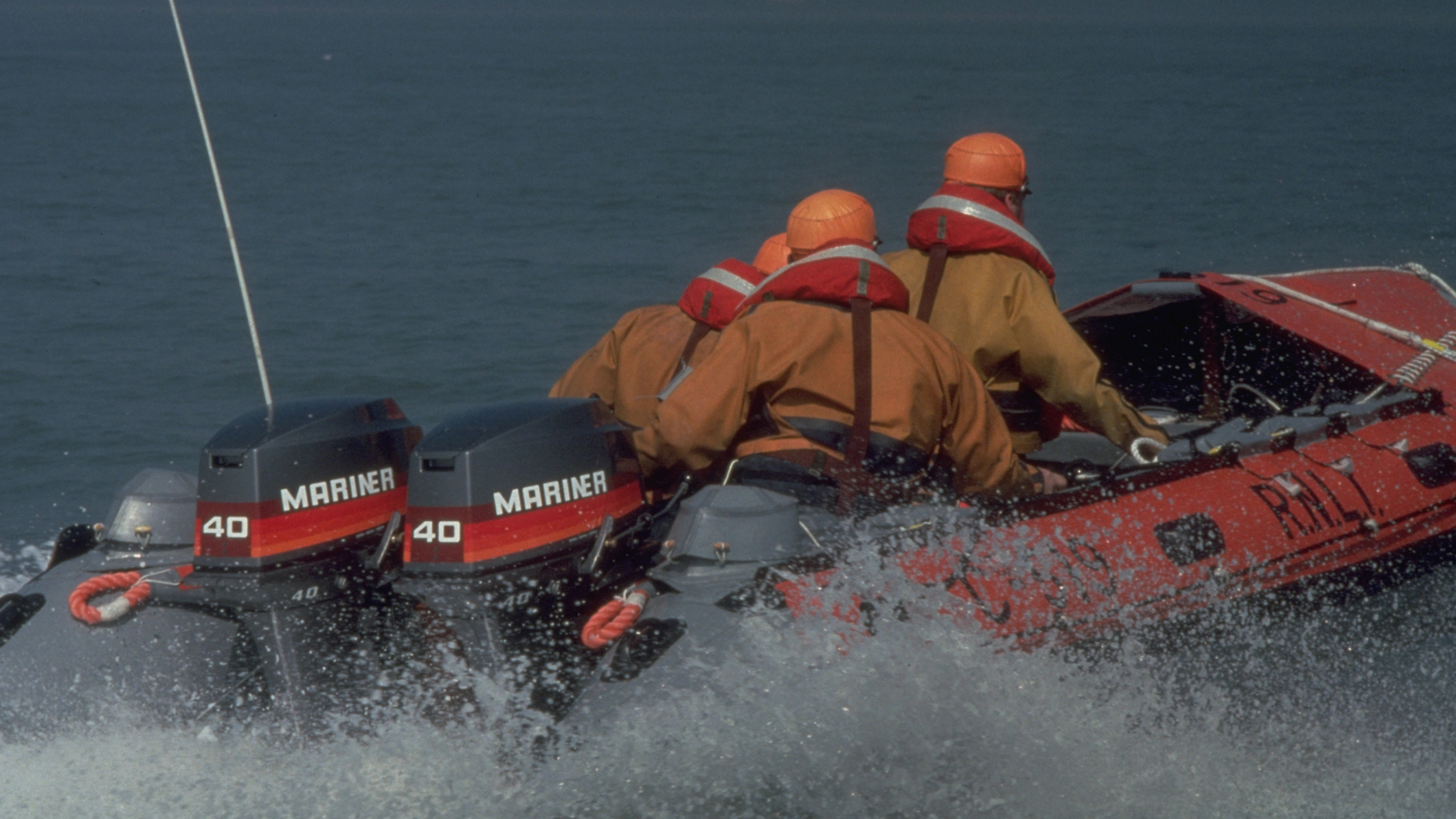 A stern view of Aberystwyth’s C class lifeboat, with three crew onboard. They’re wearing bright orange helmets, darker orange lifejackets and light orange crew kit.