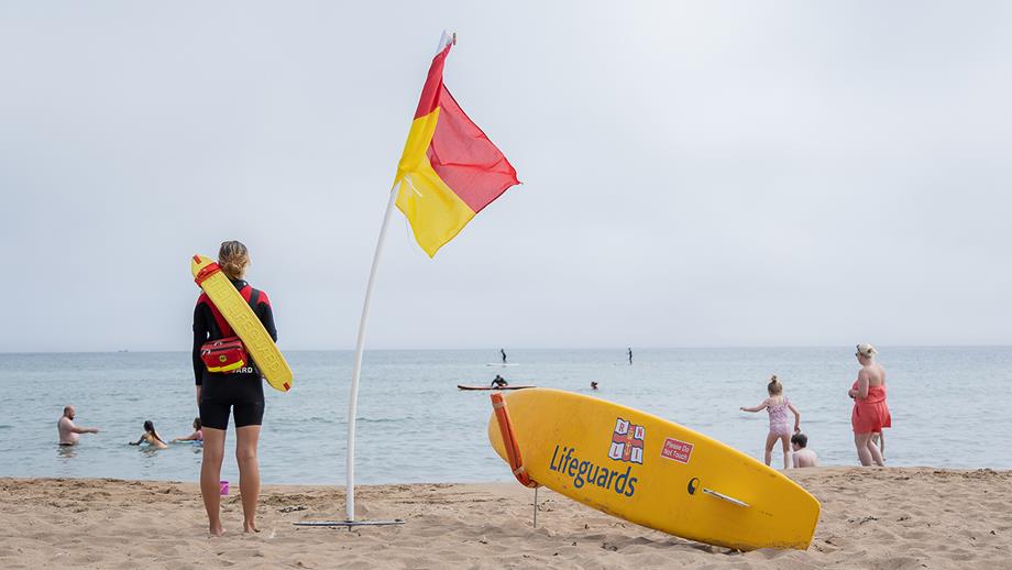A lifeguard stands facing the sea, watching over it. Next to her is a rescue board and a red and yellow flag.