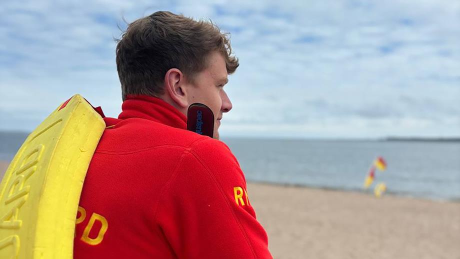 RNLI Lifeguard Callum McDonald, a white man with light brown hair, looks out to sea past red and yellow flags in the distance. He is wearing a red lifeguard fleece and carries a yellow rescue float over his shoulder.
